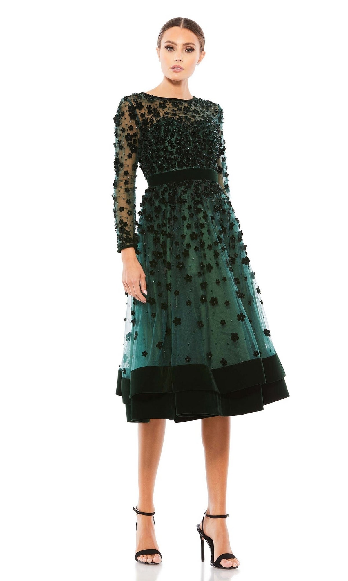 A woman wearing the Mac Duggal 67007 knee-length wedding-guest dress in dark green with black floral embellishments and sheer details, paired with black high-heeled sandals.